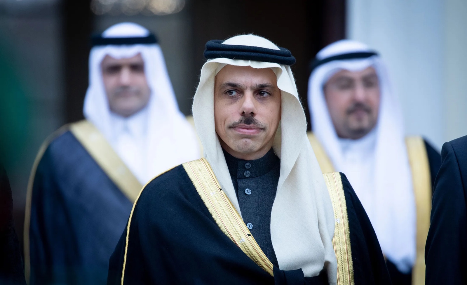 Faisal bin Farhan Al-Saud in a white ghutra and black bisht, with two Saudi officials out of focus behind him, 2019