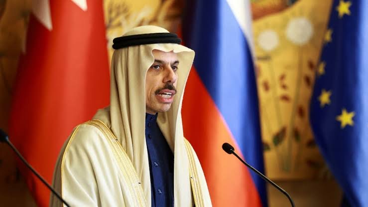 Faisal bin Farhan Al-Saud speaking at a lectern between two microphones, with several national flags and the flag of the European Union behind him