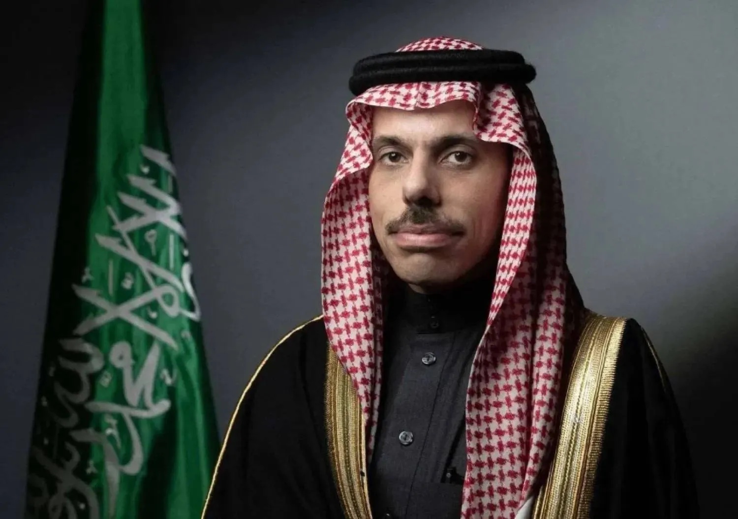 Formal portrait of Faisal bin Farhan Al-Saud in a red-and-white shemagh and black bisht with gold trim, standing beside the Saudi flag against a grey background
