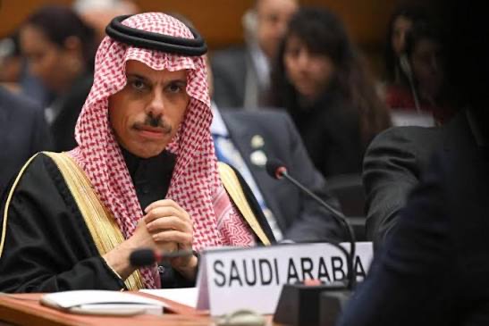 Faisal bin Farhan Al-Saud seated behind a 'Saudi Arabia' nameplate at a multilateral meeting, hands clasped